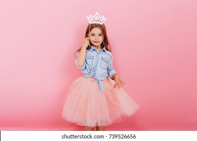Cheerful little girl with long brunette hair in tulle skirt holding princess crown on head  isolated on pink background. Celebrating brightful carnival for kids, birthday party, having fun of cute kid