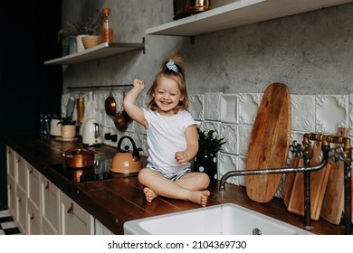 A cheerful little girl in pajamas is sitting on the kitchen cabinet playing with water from the sink washing her hands and monitoring hygiene in the kitchen at home. Kid fooling around splashing water