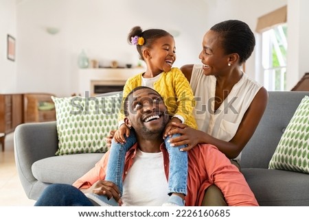 Cheerful little girl sitting on father shoulder while playing with mother at home. Happy black  family enjoying weekend at home. Cute little daughter sitting on fathers shoulder and play with her mom.