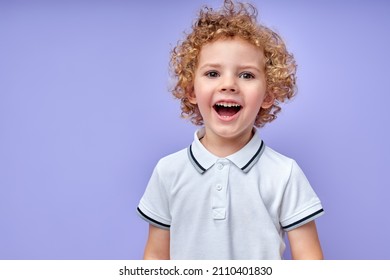 Cheerful Little Kid With Curly Hair Laughing After Hearing Funny Joke From Friend, Child Is Looking At Camera, Wearing Casual T-shirt, Isolated Over Purple Studio Background. Portrait. Human Emotions