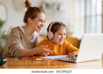 Cheerful Little Schoolgirl In Headphones Talking To The Teacher While Studying Remotely Via Laptop At Home With Happy Mom Sitting Nearby And Giving Support