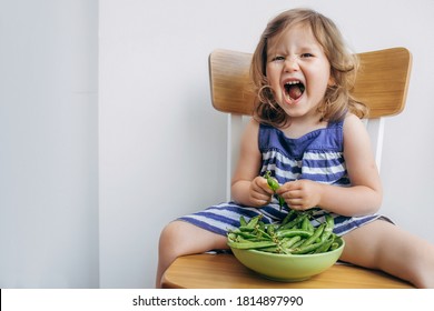 Cheerful Little Todler Girl In A Striped Dress Eating Peas And Grimacing