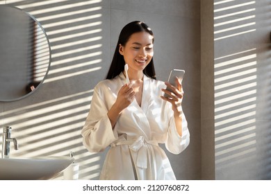 Cheerful Long-haired Millennial Korean Lady In Silk Bathrobe Using Mobile Phone While Doing Morning Routine In Bathroom, Brushing Teeth, Looking At Smartphone Screen And Smiling, Sun Flare