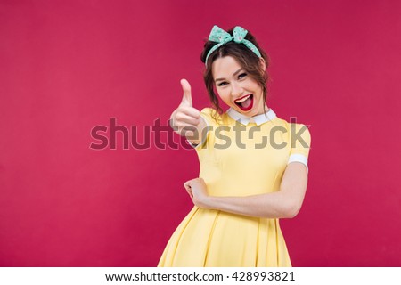 Cheerful lovely young woman in yellow dress showing thumbs up over pink background