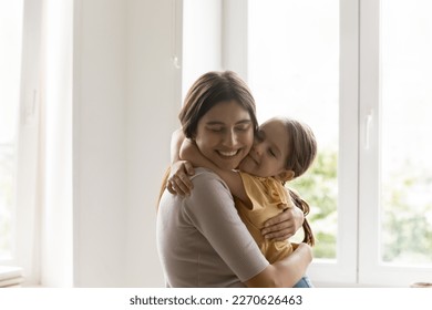Alegre hija de amor abrazando y besando a mamá feliz. Madre positiva sosteniendo a una niña en brazos, apretando, acurrucando a un niño con cuidado, ternura, de pie contra la ventana, fondo blanco