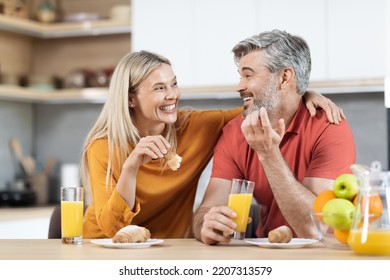 Cheerful Loving Middle Aged Man And Woman Sitting At Kitchen Table, Having Snack, Talking At Home, Married Couple Enjoying Time Together, Cuddling, Talking, Eating Pastry, Drinking Juice, Copy Space