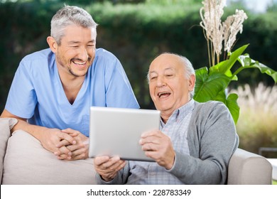 Cheerful Male Caretaker And Senior Man Using Tablet PC At Nursing Home Porch