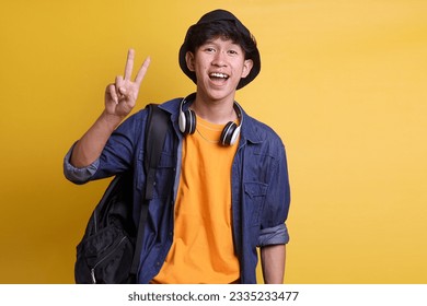 Cheerful male college student showing two fingers v-sign peace greeting gesture in casual clothes, headphones around his neck and wear a bucket hat 
