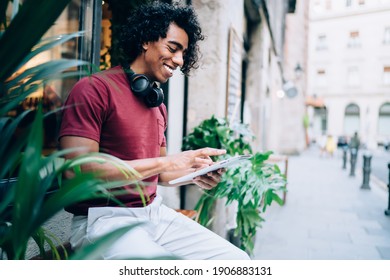 Cheerful male connecting to 4g wireless on modern touch pad for making text chatting in social media, smiling hipster guy reading travel publication resting at sidewalk during daytime in city