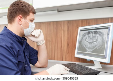 Cheerful male dentist is sitting at the table and using computer. He is analyzing x-ray of human teeth. The man is looking at the screen seriously. He is wearing gloves and mask