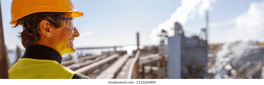 Cheerful Male Engineer In Work Vest Looking Away And Smiling While Standing On Territory Of Manufacturing Plant