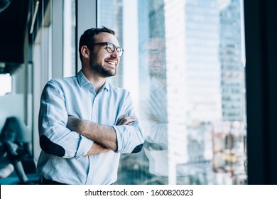 Cheerful male entrepreneur with crossed hands standing near office window view and smiling during work day in company, Caucasian successful corporate boss feeling good from wealthy lifestyle - Shutterstock ID 1600820323