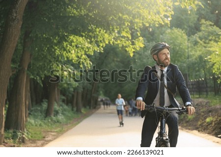 Cheerful male entrepreneur in suit, riding on bicycle after workday in city park. Front view of smiling man in smart casual enjoying trip, while driving bike in warm day. Concept of lifestyle.