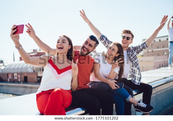 Cheerful male and female best friends enjoying\
together pastime at roof house making selfie pictures with city on\
background, joyful hipster guys shooting funny video during double\
date gathering