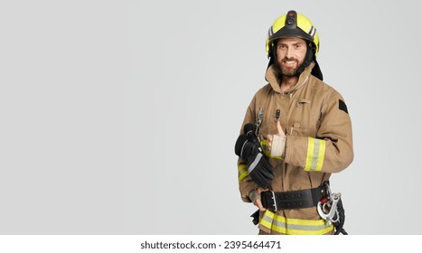 Cheerful male firefighter in uniform gesturing with thumb up. Front view of smiling bearded fireman giving like, while looking at camera, isolated on gray, copy space. Concept of work, hand gesture.
