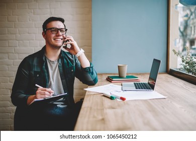Cheerful male freelancer making telephone call share good news about project working in cafe interior,happy hipster guy having smartphone conversation while studying in good mood writing in planner