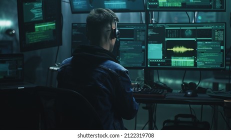 Cheerful male hacker in headset rejoicing in a successful cyber attack in a dim room of cybercriminals with computer equipment and software