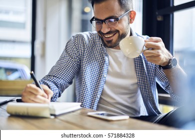 Cheerful male journalist in trendy glasses happy to finishing work on book review rereading written in notebook text before calling to editor to present publication drinking coffee for lunch in cafe - Powered by Shutterstock