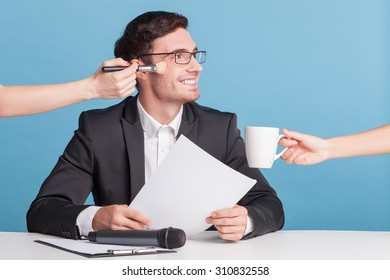 Cheerful Male Newscaster Is Resting On A Break. He Is Sitting At The Desk And Smiling. The Female Hand Is Giving A Cup Of Coffee To Him And Another Arm Is Doing Him Make-up. Isolated 