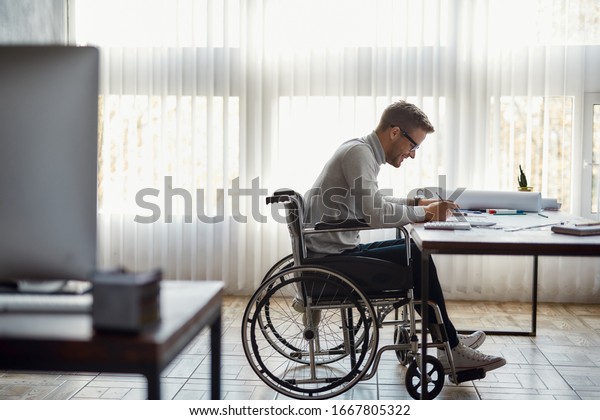 Cheerful\
male office worker in a wheelchair drawing something and smiling\
while working at his workplace in the modern office. Disability and\
handicap concept. Construction and\
architecture