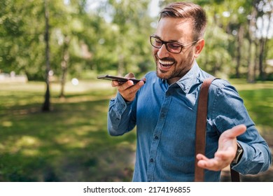 Cheerful Male Professional Talking On Smart Phone Speaker. Young Businessman Wearing Formals Is Carrying Laptop Bag. He Is Walking Against Trees At Public Park.