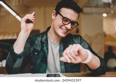 Cheerful male student satisfied with useful apps on digital smartwatch looking at touchscreen, smiling hipster guy managing time using organizer on wearable computer looking at display in cafe