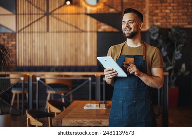 A cheerful male waiter in a stylish apron stands with a digital tablet, ready to take orders in a contemporary restaurant setting. - Powered by Shutterstock