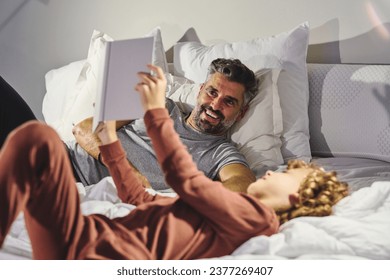 Cheerful man and boy in sleepwear having fun while lying on soft bed and reading interesting book together in cozy bedroom at home