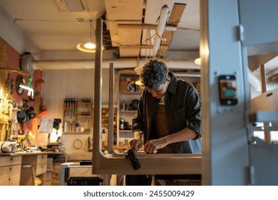 Cheerful man carpenter working with wooden handmade table. Interested craftsman pulls up bolts in frame of table screws to tabletop. Joiner feels satisfaction happiness gladness from work process. - Powered by Shutterstock