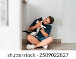 A cheerful man in casual clothes sits on the floor, affectionately holding his black and white cat.