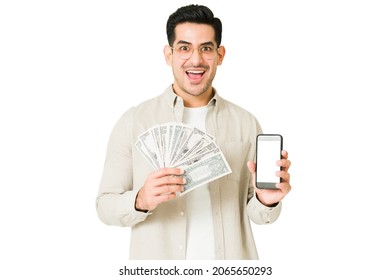 Cheerful Man Earning A Lot Of Money With His Online Work. Excited Man Holding His Smartphone And A Lot Of Money