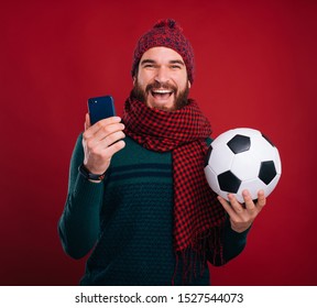 Cheerful Man Is Holding His Phone And A Soccer Ball On Red Background.