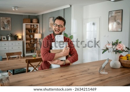 Cheerful man holding a stack of parcels in a modern apartment, radiating satisfaction from recent online purchases. Enjoying the convenience of ecommerce and delivery at home