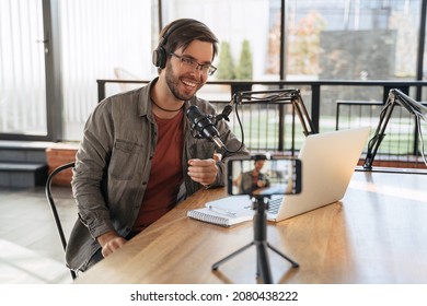 Cheerful man host with stubble laughing and gesticulating while streaming video podcast in broadcasting studio, using microphone, smartphone and laptop. Famous vlogger shooting video for his channel - Powered by Shutterstock