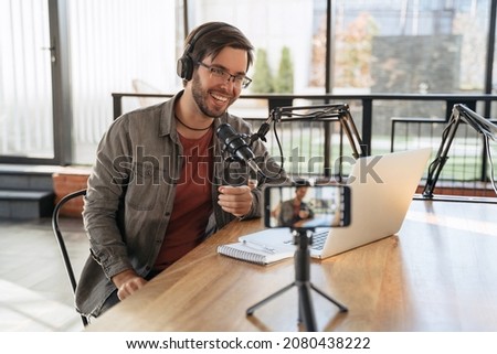 Cheerful man host with stubble laughing and gesticulating while streaming video podcast in broadcasting studio, using microphone, smartphone and laptop. Famous vlogger shooting video for his channel