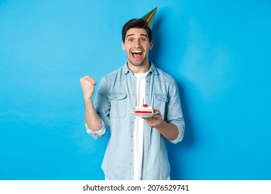 Cheerful man looking happy, celebrating birthday in party hat, holding b-day cake and make fist pump, standing over blue background