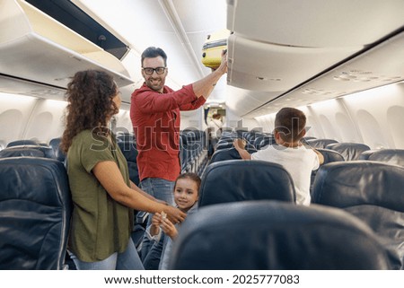 Cheerful man looking at his wife with a smile and putting carry on luggage in compartment while traveling together with his family by plane. Family vacation, transportation concept