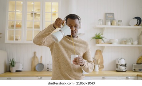 Cheerful Man Pours Hot Tea Into Cup From Kettle Dancing Against Countertop In Kitchen. African American Guy Listens To Dynamic Music In Headphones