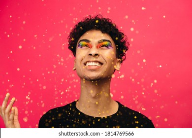 Cheerful man with rainbow eye makeup throwing up the glitter. Excited gender fluid male looking at falling glitters against red background.