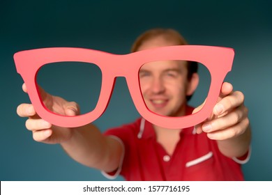 Cheerful man in a red shirt holding big red glasses and looking through . Good Vision Concept. Approach to exaggeration and distortion of facts