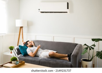 Cheerful man relaxing on the sofa and using the remote to turn on the ac unit during a hot summer