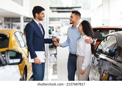 Cheerful Man Sales Associate Giving Happy Young Middle Eastern Couple Key From Brand New Car And Smiling, Side View. Beautiful Arabic Family Buying Brand New Auto At Luxury Dealership Salon