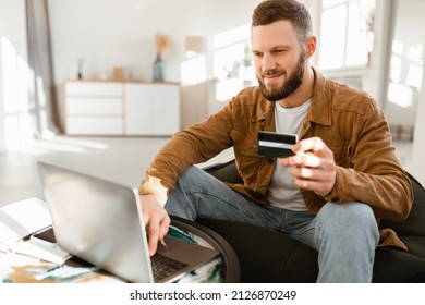 Cheerful Man Shopping Online Using Laptop Computer And Credit Card Making Payment Sitting At Home. Male Customer Paying Via Internet Banking. Modern Consumerism And Ecommerce