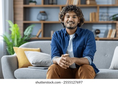 A cheerful man sits comfortably on a plush sofa, exuding warmth and friendliness. The home interior setting is modern, with soft furnishings contributing to a cozy atmosphere. - Powered by Shutterstock