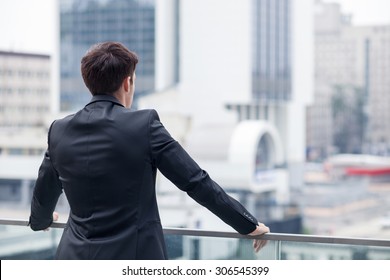 Cheerful man in suit is enjoying the view of the city from a balcony of his office. He is standing and relaxing. Focus on his back. Copy space in right side