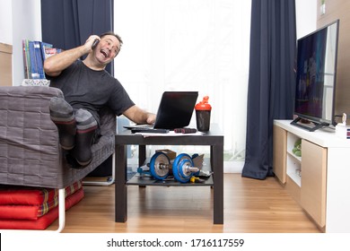 Cheerful Man Talking On The Phone While Sitting In Living Room With TV And Laptop On A Table. Home Office.