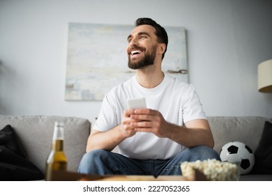 cheerful man using smartphone near bottle of beer and tasty food on blurred foreground