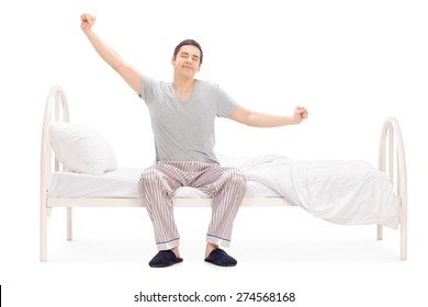 Cheerful man waking up from sleep and stretching seated on his bed isolated on white background
