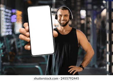 A cheerful man wearing headphones and a black tank top is standing in a gym, extending his arm to display a blank screen on his smartphone - Powered by Shutterstock