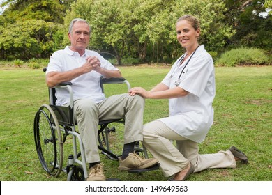 Cheerful Man In A Wheelchair With His Nurse Kneeling Beside Smiling At Camera In The Park